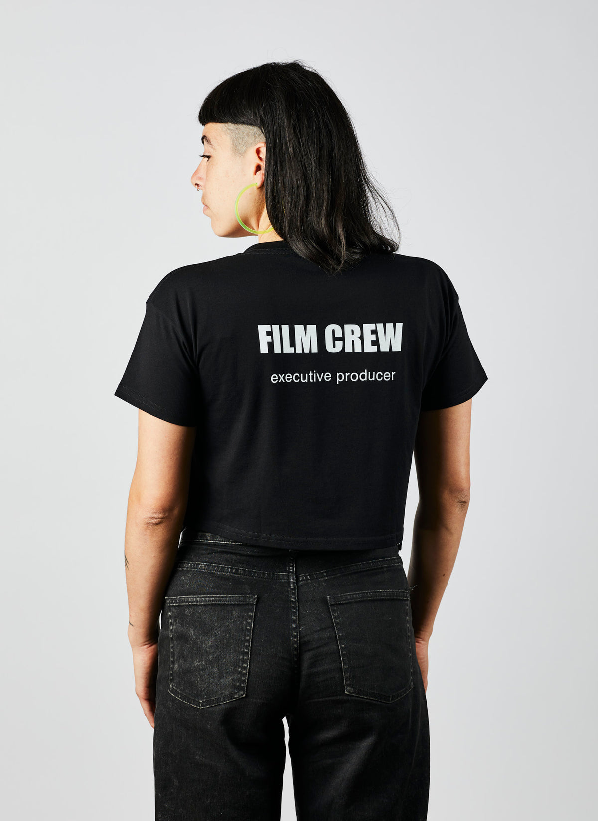 Reverse of a woman wearing a black, cropped &quot;Executive Producer&quot; shirt in a white studio