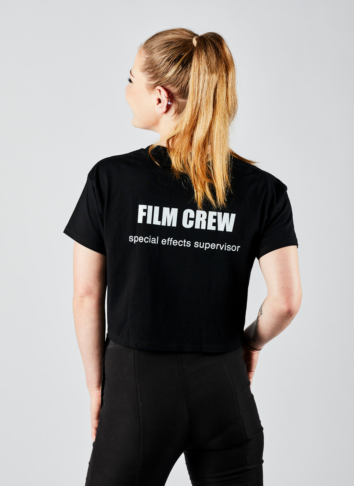Reverse angle of a long-haired woman wearing a cropped black film crew shirt with &quot;Special Effects Supervisor&quot; printed on the back